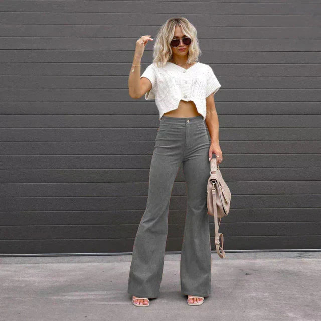 Vrouw in grijze flared broek en witte crop top voor grijze muur, draagt zonnebril en houdt beige tas. Modieuze dameskleding, casual stijl.