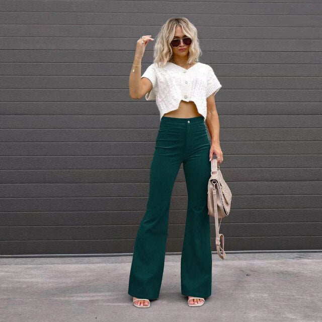 Vrouw in witte crop top en groene flared broek poseert voor grijze muur, draagt zonnebril en beige tas. Modieuze dameskleding, trendy outfit.